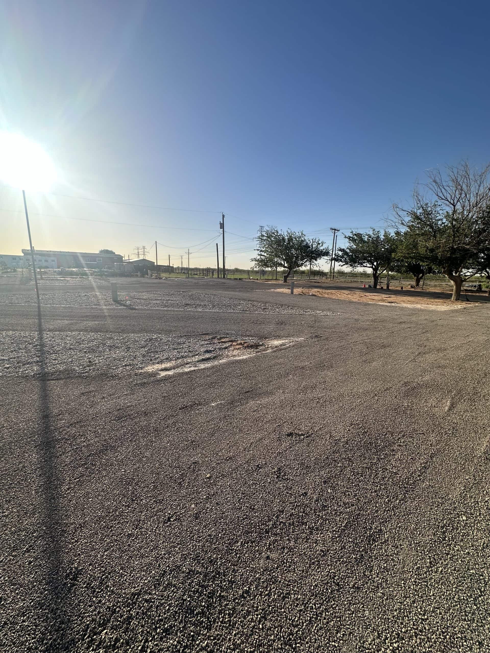 A wide gravel lot under a bright, clear sky with the sun low on the horizon. Sparse trees line one side, and utility poles are visible in the distance, casting long shadows across the ground.