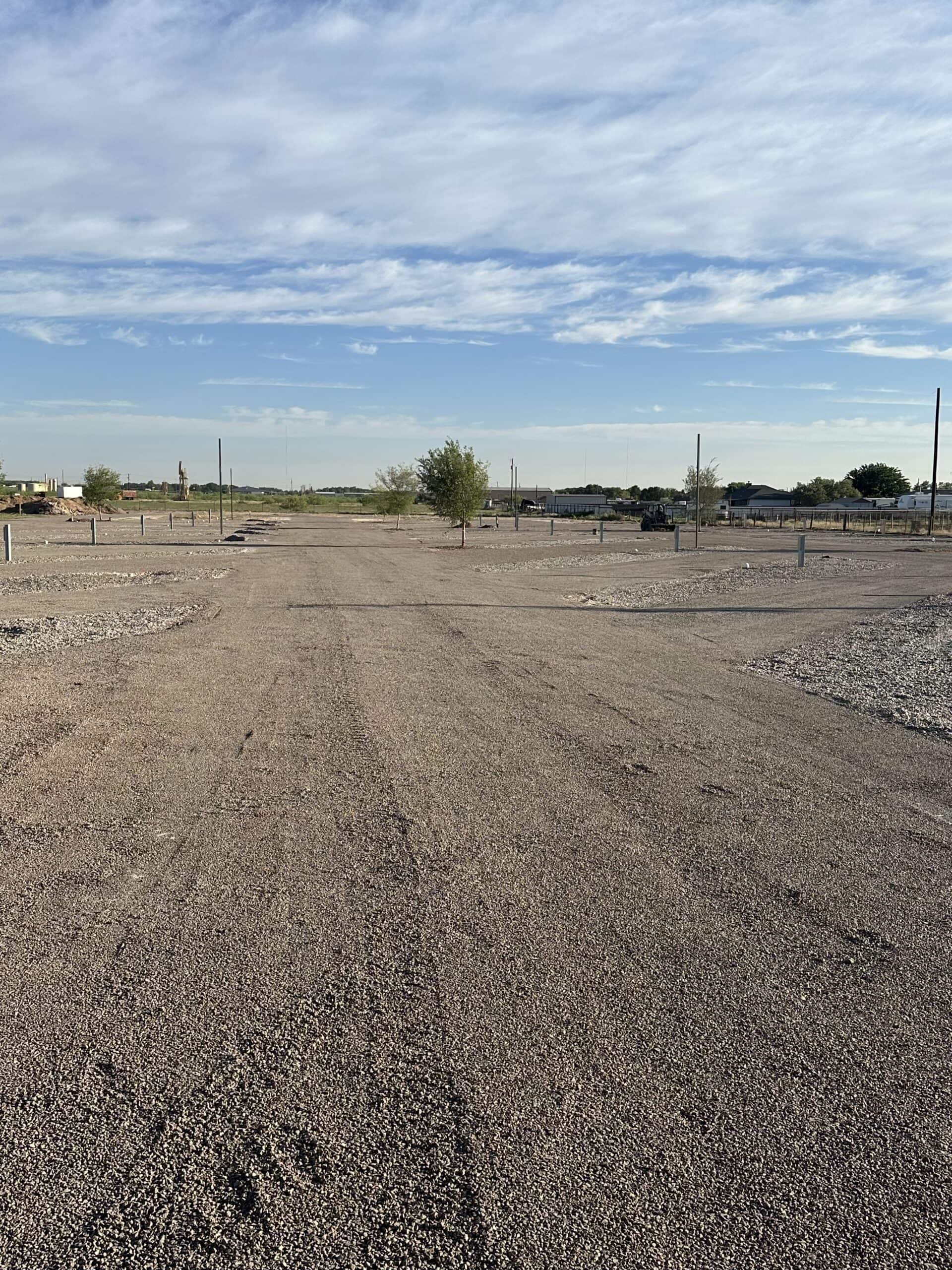 A barren, gravel-covered landscape under a blue sky with scattered clouds. Sparse vegetation is scattered across the flat terrain, with a few small trees and utility poles in the distance.
