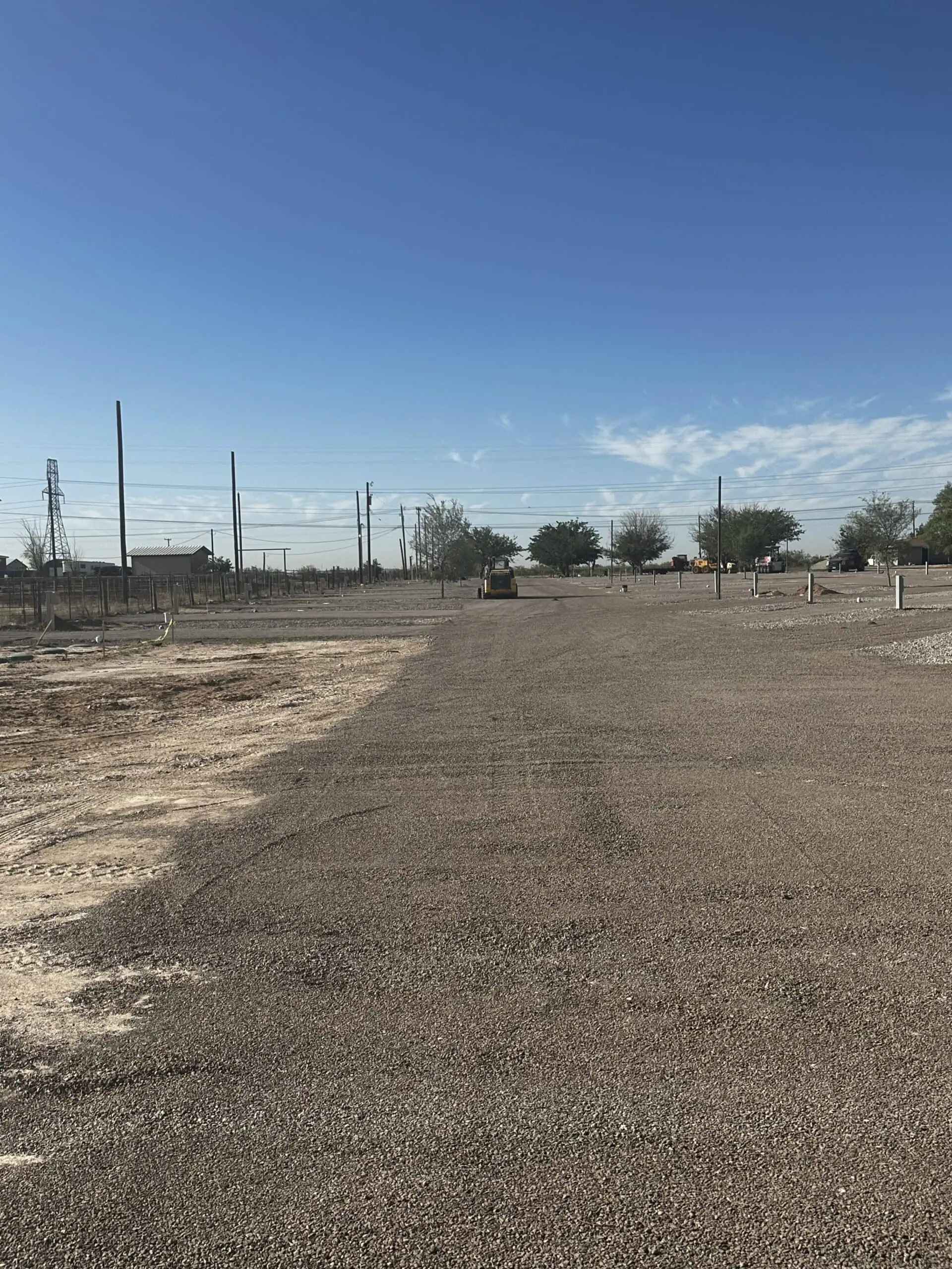 A construction site with a gravel-covered area under a clear blue sky. In the background, trees, utility poles, and a small piece of construction machinery are visible. The landscape appears flat and expansive.